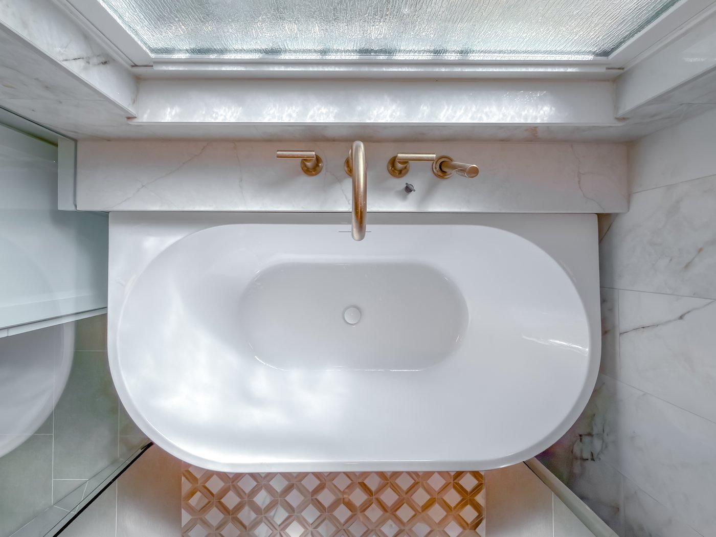The same bathroom after renovation, with a freestanding soaking tub, marble-veined tile and a patterned mosaic floor