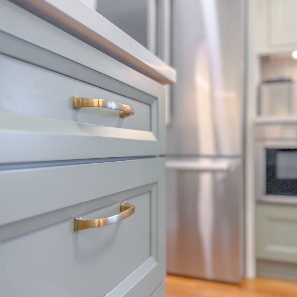 Close detail of soft sage inset cabinet drawers with aged brass bow pulls beside a stainless refrigerator. Kitchen renovation in Tallahassee, FL