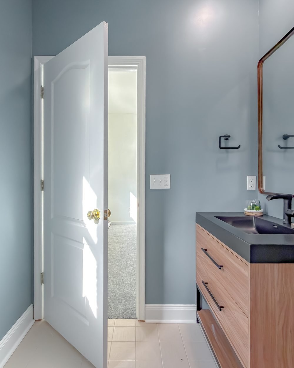 Bathroom with a natural oak floating vanity, black composite basin and bronze-framed mirror against pale blue walls. Bathroom renovation in Tallahassee, FL
