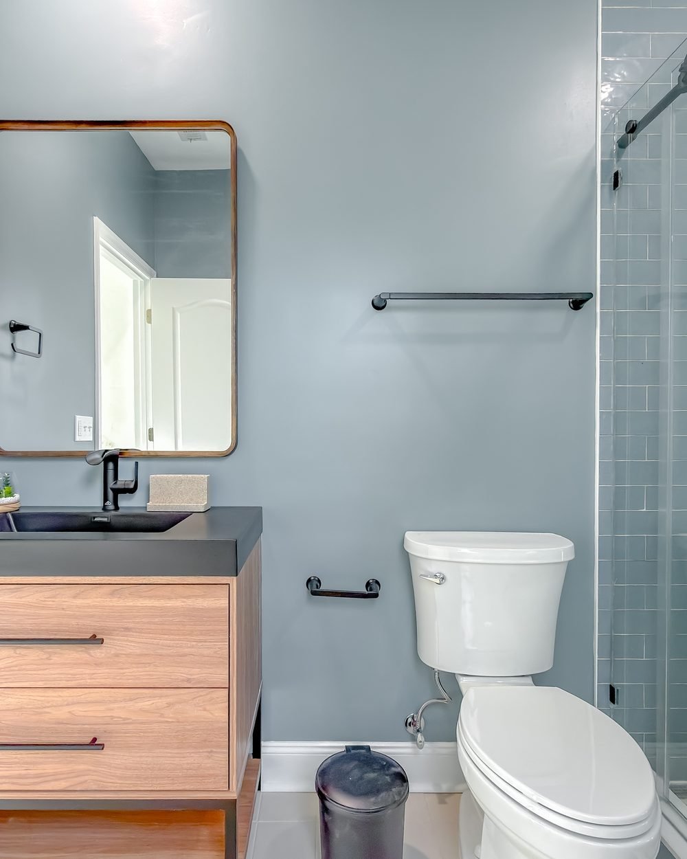 Bathroom with an oak floating vanity, bronze-framed mirror and matte black fittings against pale blue walls. Tallahassee, FL